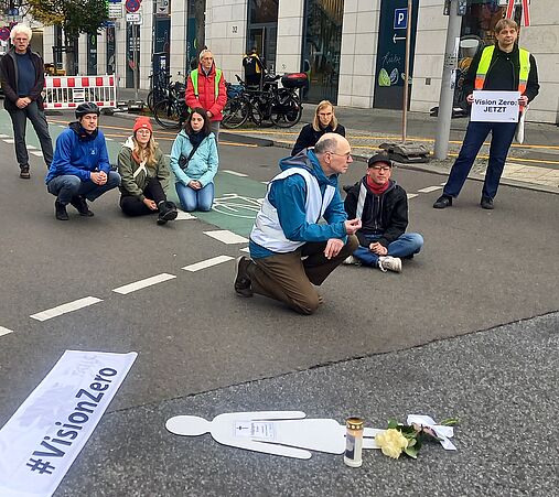 mehere Menschen, teils sitzend, teils knieend auf der Straße. Im Vordergrund liegt eine weiße Symbolfigur